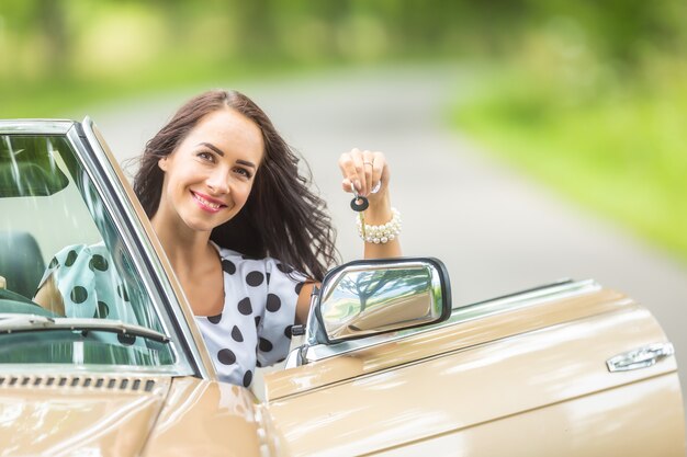 Une femme sourit en tenant des clés de voiture dans sa main, assise dans une voiture ancienne décapotable.