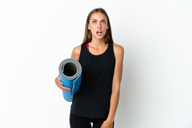 Femme de sport allant à des cours de yoga tout en tenant un tapis sur fond blanc isolé à la recherche et avec une expression surprise