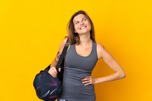 Femme de sport slovaque avec sac de sport isolé sur fond jaune posant avec les bras à la hanche et souriant