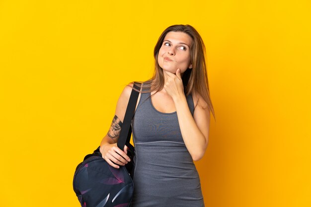 Femme de sport slovaque avec sac de sport isolé sur mur jaune ayant des doutes et de la pensée