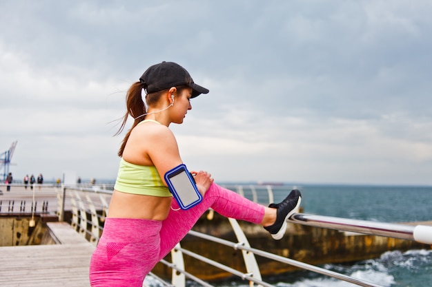 Femme de sport en tenue de sport avec un casque dans les oreilles faisant étirement de la jambe avant de s'entraîner sur la plage en plein air