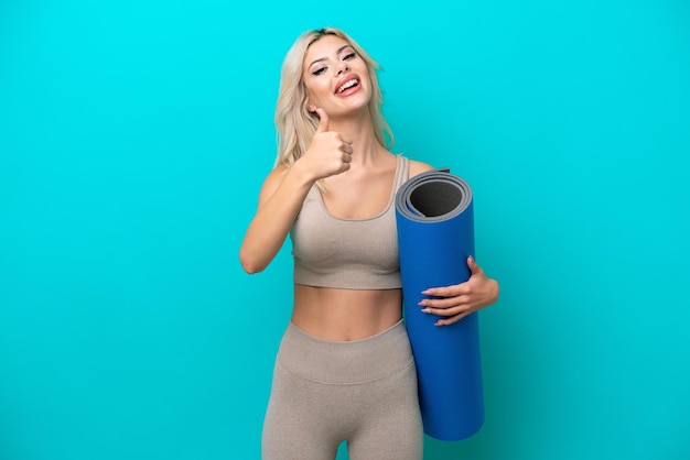 Femme sportive allant à des cours de yoga tout en tenant un tapis isolé sur fond bleu avec les pouces vers le haut parce que quelque chose de bien s'est produit
