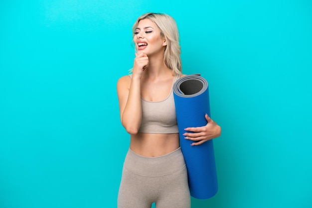 Femme sportive allant à des cours de yoga tout en tenant un tapis isolé sur fond bleu regardant sur le côté et souriant