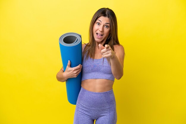 Femme sportive allant à des cours de yoga tout en tenant un tapis isolé sur fond jaune non surpris et pointant vers l'avant