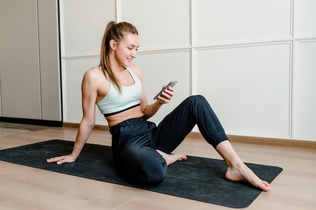 Femme sportive assise sur un tapis de yoga à la maison avec téléphone en ligne