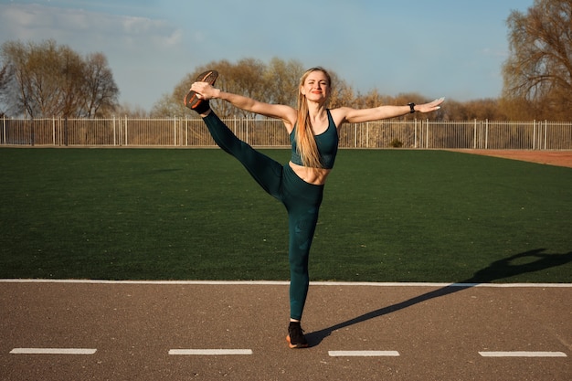 Une femme sportive athlétique s'étire la jambe en faisant des exercices de fitness à l'extérieur sur le terrain de sport