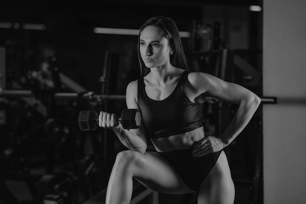 Une femme sportive fait des boucles d'haltères avec son coude sur un genou dans une salle de sport. Une fille brune musclée porte un haut noir et un short court taille haute.