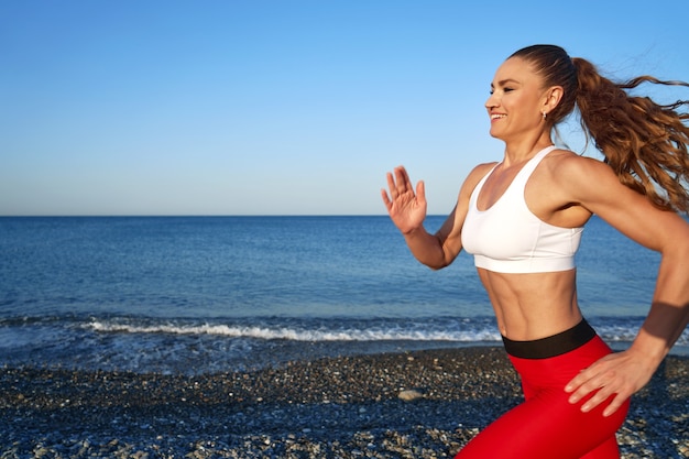 Femme sportive positive sur un matin d'été jogging sur la plage en leggings rouges sur le fond de la côte de la mer près de uo