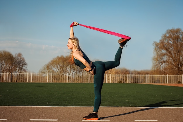 Une femme sportive s'étire le dos et la jambe à l'aide d'un extenseur, d'une bande de résistance à l'extérieur du stade.