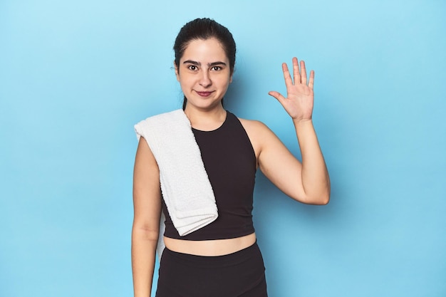 Femme sportive avec une serviette sur un studio bleu souriant joyeux montrant le numéro cinq avec les doigts