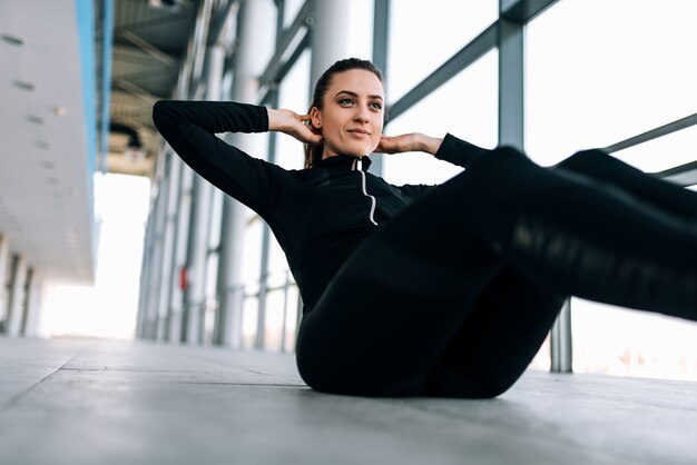 Femme sportive en survêtement noir faisant des sit-ups à l&#39;intérieur.