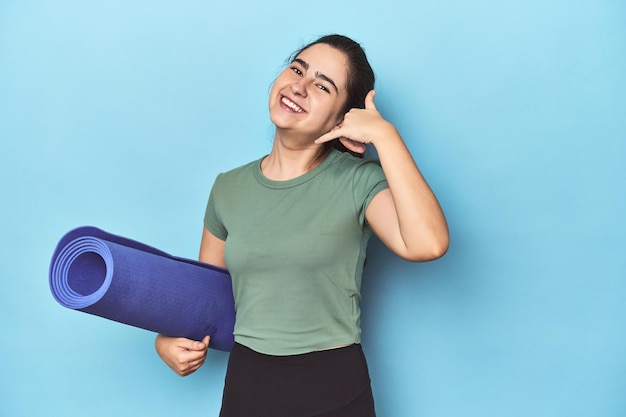 Femme sportive avec tapis de yoga sur bleu montrant un geste d'appel de téléphone portable avec les doigts