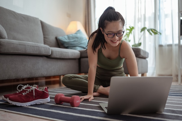 Une femme sportive en tenue de sport est assise sur le sol avec des haltères et utilise un ordinateur portable à la maison