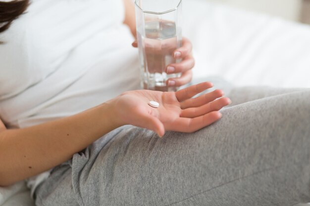 Femme stressée buvant une pilule ou un médicament avec un verre d'eau sur le lit à la maison