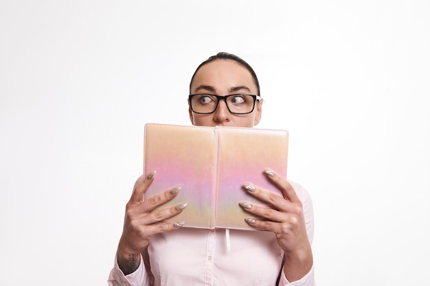 Femme stupéfaite et surprise en détournant les yeux et couvrant sa bouche avec un livre rose. Isolé sur fond blanc avec espace copie