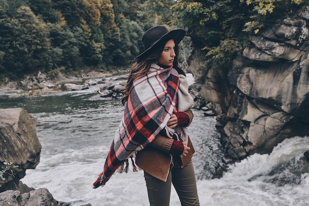 Femme avec style. Jolie jeune femme recouverte d'une couverture en détournant les yeux tout en se tenant près de la rivière dans les montagnes