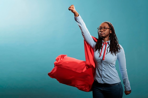 Photo femme de super-héros afro-américaine puissante et courageuse se faisant passer pour un défenseur de la justice tout en agissant comme voler sur fond bleu. jeune adulte fort portant une cape de héros rouge. tourné en studio.