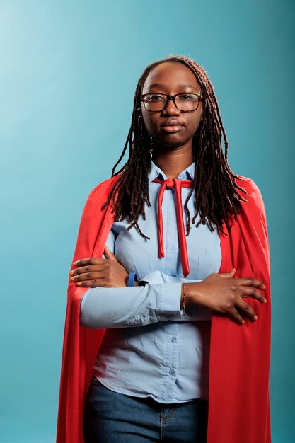 Femme de super-héros confiante et courageuse debout sur fond bleu avec les bras croisés tout en regardant la caméra. Défenseur de la justice avec des capacités de superpuissance portant une puissante cape rouge. Tourné en studio.