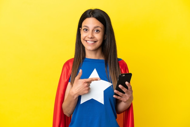 Femme de super héros isolée sur fond jaune à l'aide d'un téléphone portable et le pointant