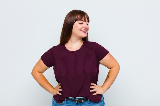 Femme en surpoids à la recherche heureuse, joyeuse et confiante, souriant fièrement et à la recherche de l'autre avec les deux mains sur les hanches