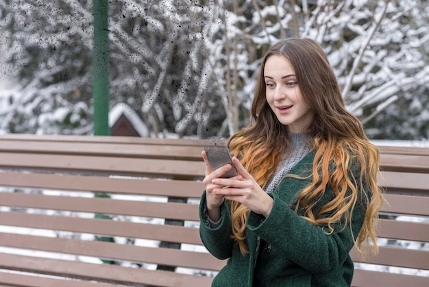 Femme surprise assise sur un banc dans un parc d'hiver et tenant un smartphone en train de se désintégrer et de disparaître