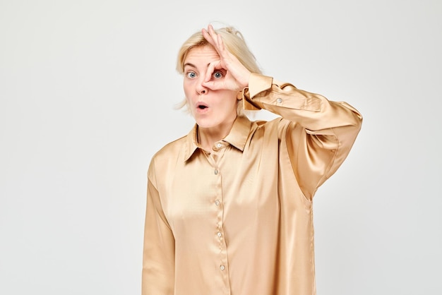 Une femme surprise en blouse beige fait un geste OK sur un fond blanc des yeux