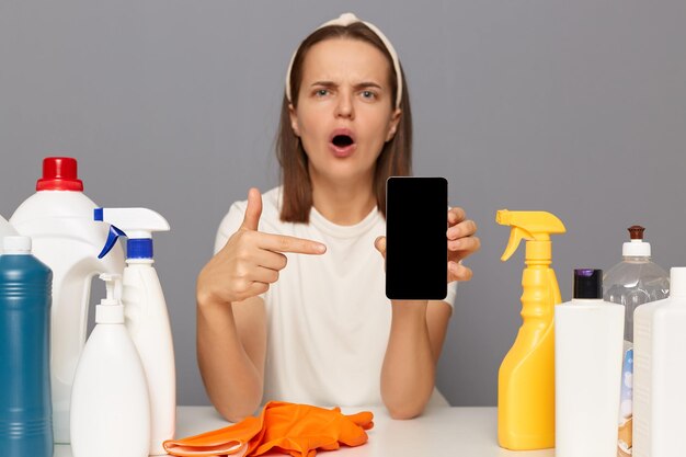 Photo femme surprise étonnée choquée posant sur le lieu de travail avec des détergents de nettoyage isolés sur fond gris pointant vers un espace vide d'affichage vide pour la publicité