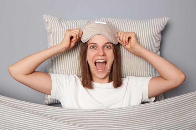 Une femme surprise et ravie en T-shirt blanc et un masque pour les yeux endormi se trouve au lit sur un oreiller sous une couverture isolée sur fond gris criant avec un visage heureux