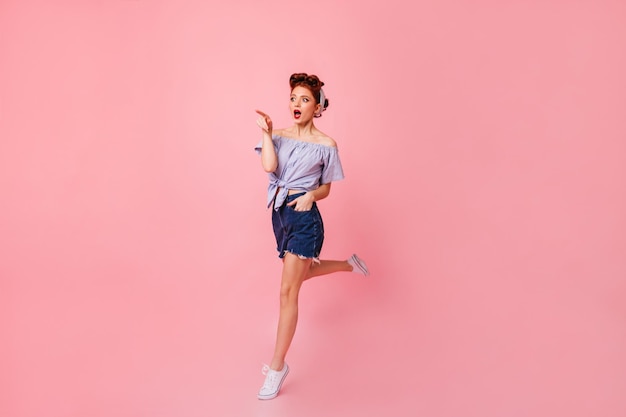 Femme surprise en short en jean pointant du doigt Dame pin-up étonnée sautant sur fond rose