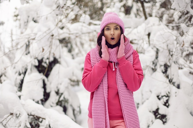 Femme surprise en vêtements roses une veste une écharpe tricotée et un chapeau se dresse dans une forêt enneigée en hiver
