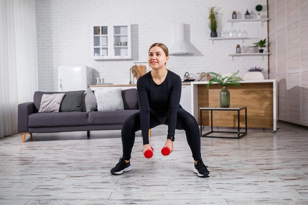 Une femme svelte fait du sport à la maison en haut et leggings. Fitness à la maison pour un beau corps. Exercices avec haltères pour le corps féminin. Squats avec haltères pour des fesses fermes