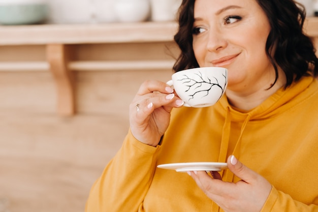 Une femme en sweat-shirt orange boit du café dans la cuisine à la maison