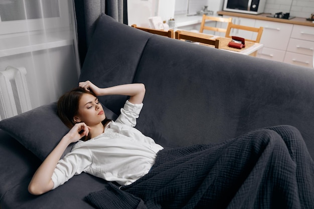 Une femme en t-shirt blanc est allongée sur le canapé sous une couverture et fait des gestes avec ses mains Photo de haute qualité