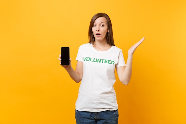 Femme en t-shirt blanc, inscription écrite, titre vert, volontaire, tenir un téléphone portable avec un écran vide vierge isolé sur fond jaune. Aide d'assistance gratuite volontaire, concept de travail de grâce de charité.