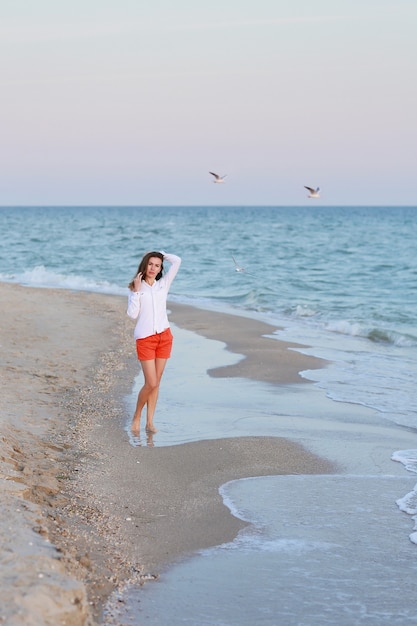 Femme en t-shirt blanc et short rouge debout près de la mer sur une plage.