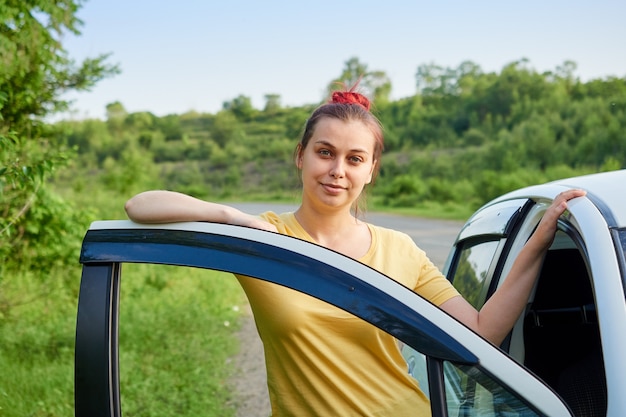 Une femme en T-shirt jaune se tient près de sa voiture sur le bord de la route en ouvrant la porte.