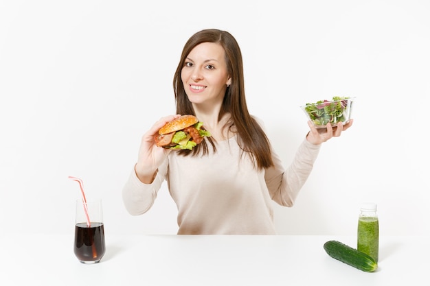 Femme à table avec des smoothies de désintoxication verts, salade dans un bol en verre, concombre, hamburger, cola en bouteille isolé sur fond blanc. Une bonne nutrition, un mode de vie sain, une restauration rapide classique, un concept de régime.