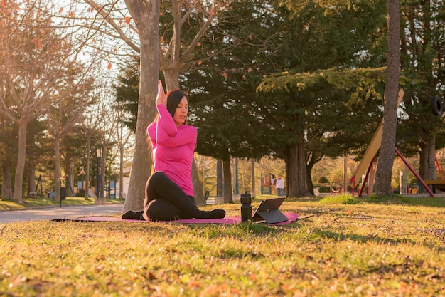 Femme avec une tablette effectuant des exercices de yoga dans un parc au coucher du soleil