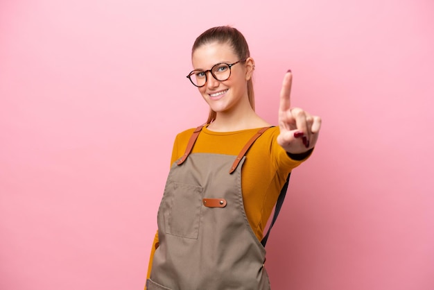 Femme avec tablier isolé sur fond rose montrant et levant un doigt