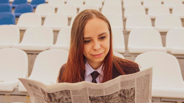 Une femme en tailleur lit un journal assise au stade