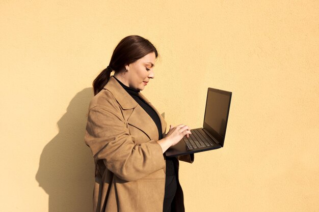 Femme tapant d'une main sur son ordinateur portable portant un manteau marron sur fond jaune et les cheveux attachés en arrière