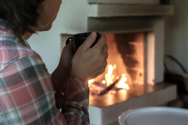 Femme avec une tasse de boisson chaude dans ses mains regarde la flamme dans une cheminée à foyer ouvert