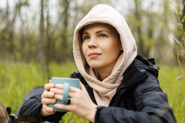 Femme avec une tasse dans la forêt, parc par temps frais sur fond vert de feuillage. Détente dans la nature, repos.