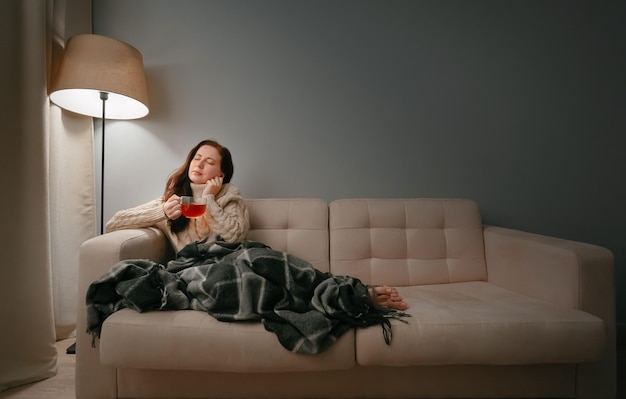 Une femme avec une tasse de thé contre la toux est assise dans une couverture sur le canapé à la maison
