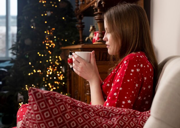 Femme avec une tasse de thé dans les mains assise sur une chaise dans une maison confortable avec un sapin pendant les vacances de Noël