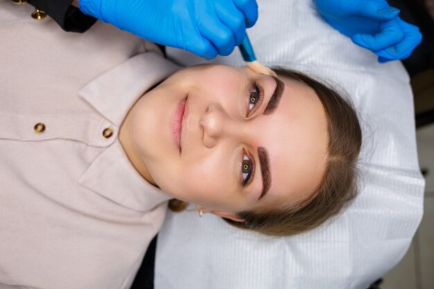 Femme avec tatouage de maquillage permanent sur ses sourcils. L'esthéticienne en gros plan fait du maquillage applique un fond de teint. Maquillage professionnel et soins cosmétiques de la peau.