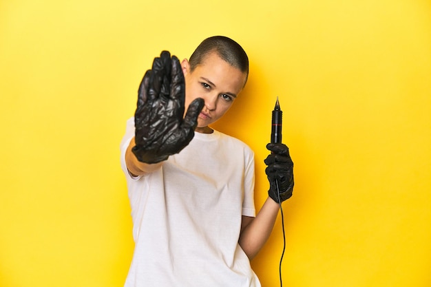 Femme tatoueuse en studio à la tête rasée à fond jaune debout avec la main tendue