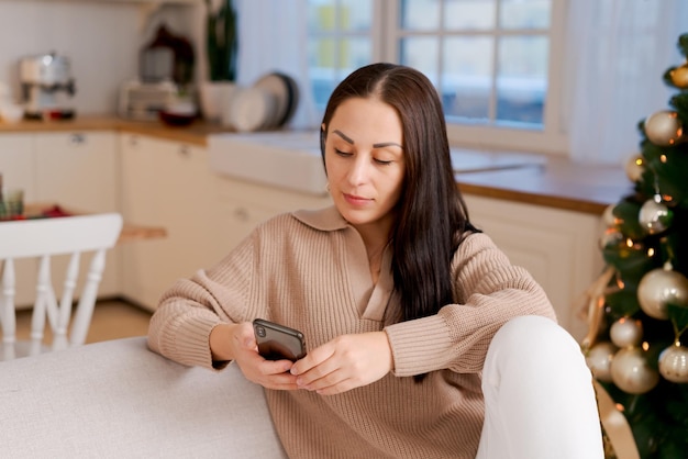 Femme avec le téléphone entre les mains assise à la maison dans la cuisine sur le canapé pendant les vacances de Noël