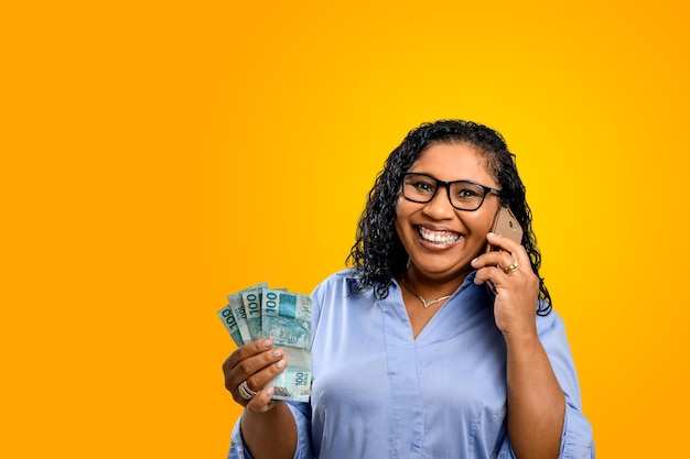 Femme tenant de l'argent parle sur téléphone portable et sourit avec contentement fond orange