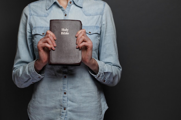 Femme tenant une Bible dans les mains sur le fond gris. Elle porte des vêtements décontractés